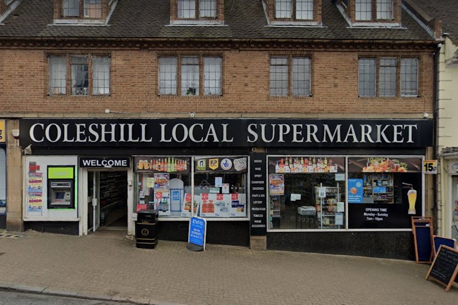 Front view of Coleshill Local Supermarket with various signs in the windows and a visible 'Welcome' sign above the entrance.