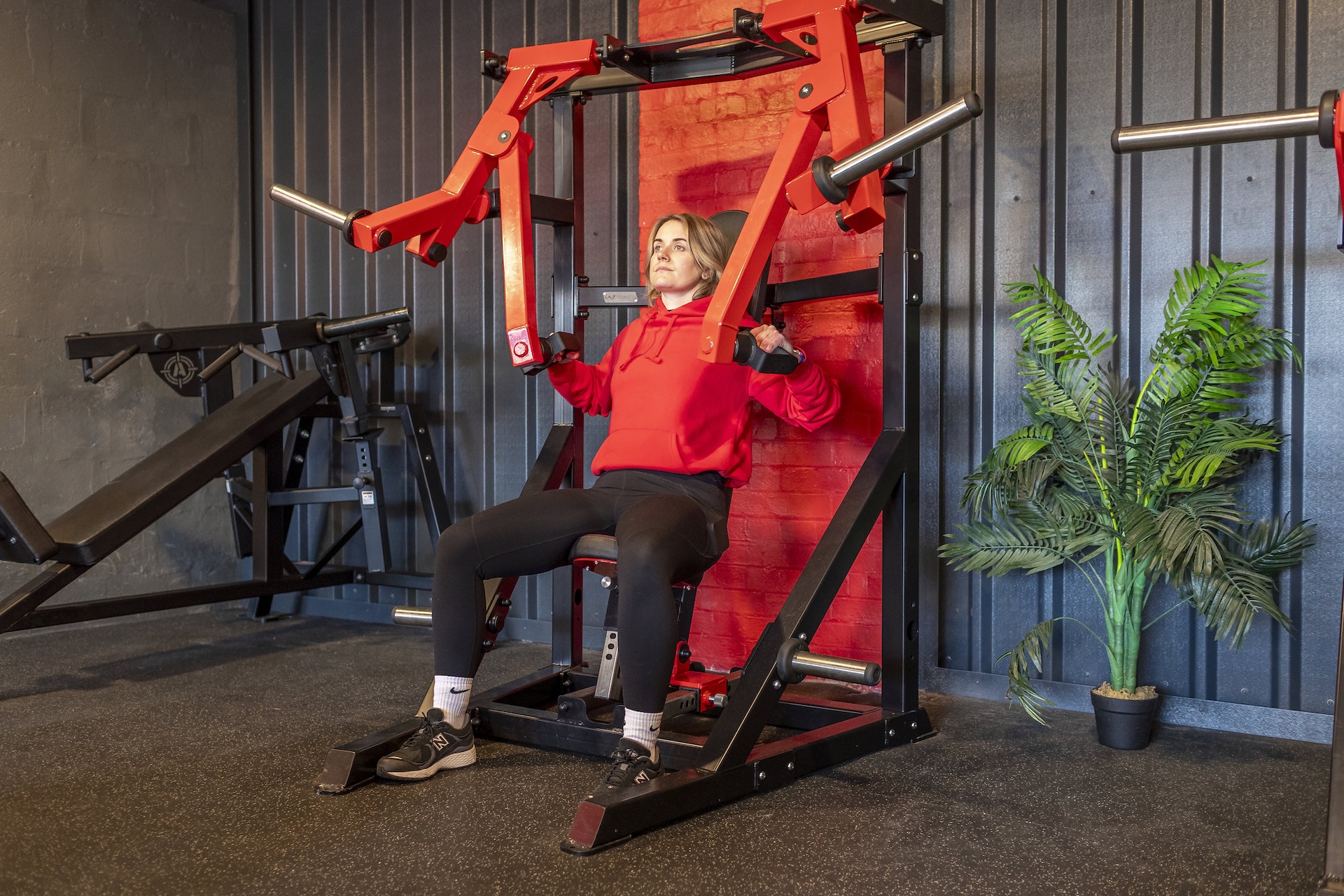 Woman in red hoodie using a shoulder press machine in a gym.
