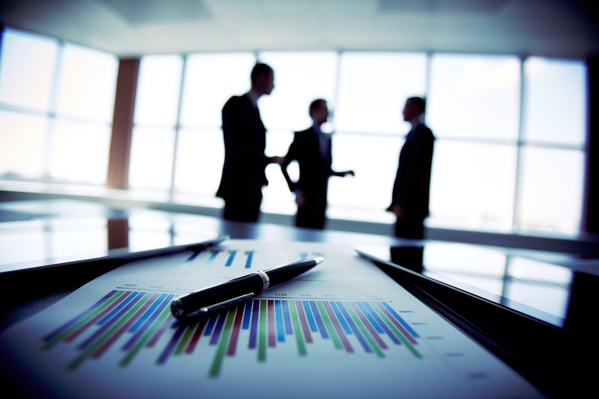Pen and colorful bar charts on paper with silhouettes of businesspeople discussing in the background.