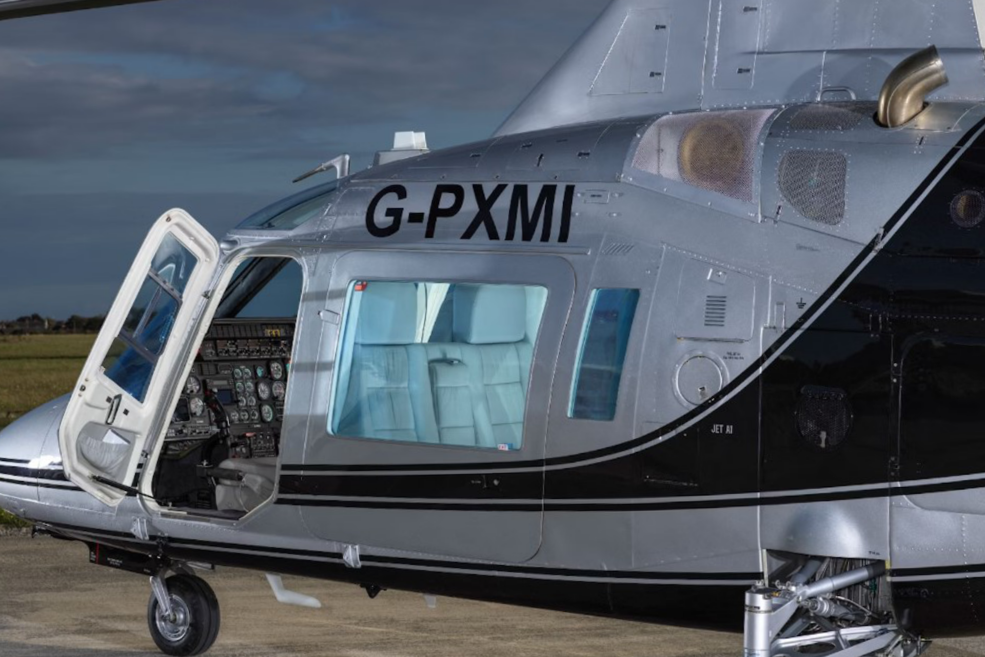 Close-up side view of a silver and black helicopter with the door open, tail number G-PXMI visible, and the cockpit instruments exposed.