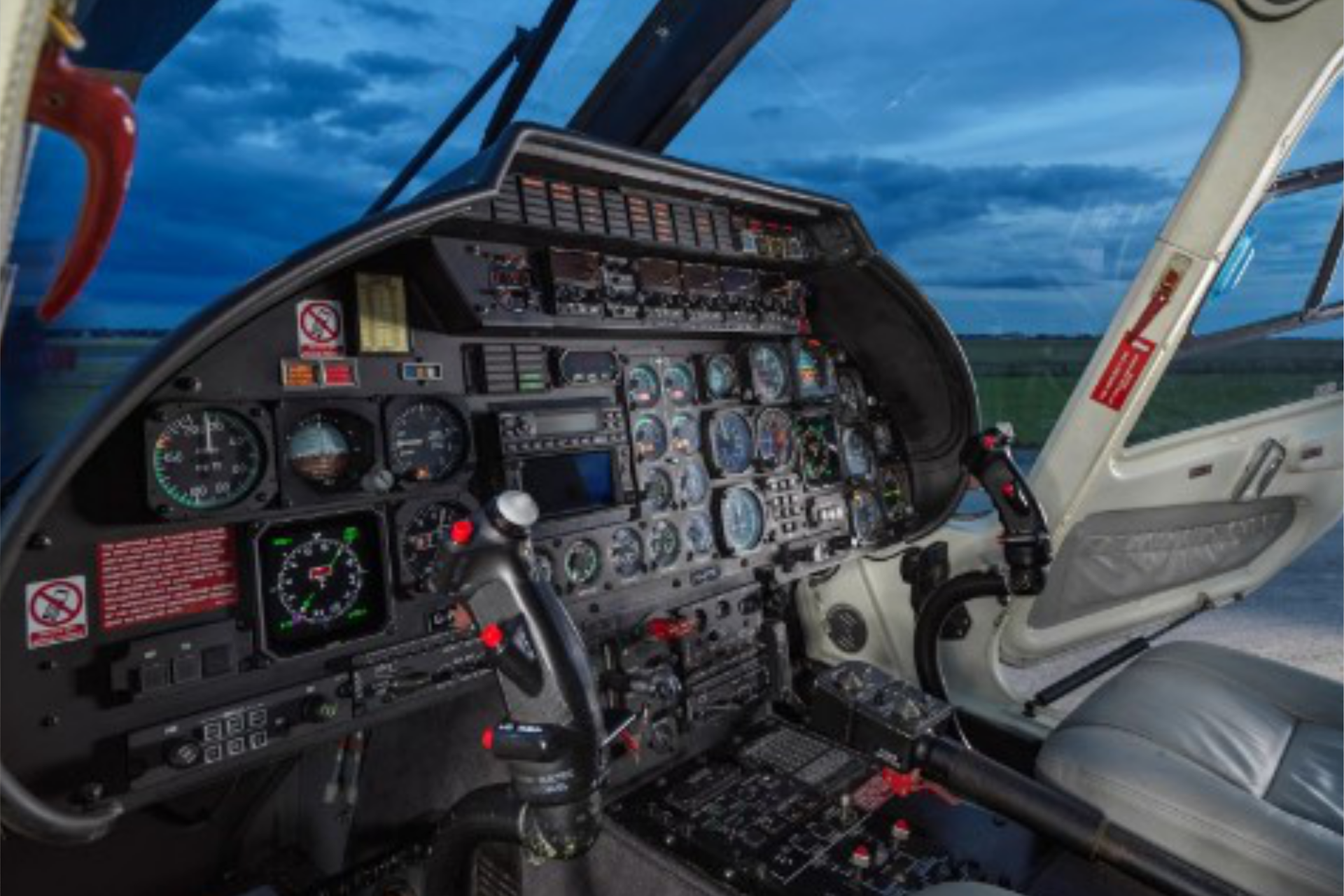 Interior view of a helicopter cockpit, showing detailed flight instruments, gauges, and control sticks, with a scenic view outside the window.